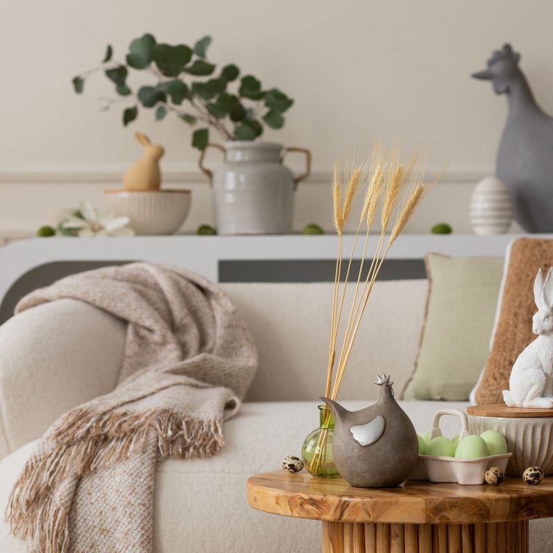 Warm and cozy composition of easter living room interior with wooden coffee table, white sofa, green easter eggs, easter bunny, vase with leaves and personal accessories. Home decor. Template.